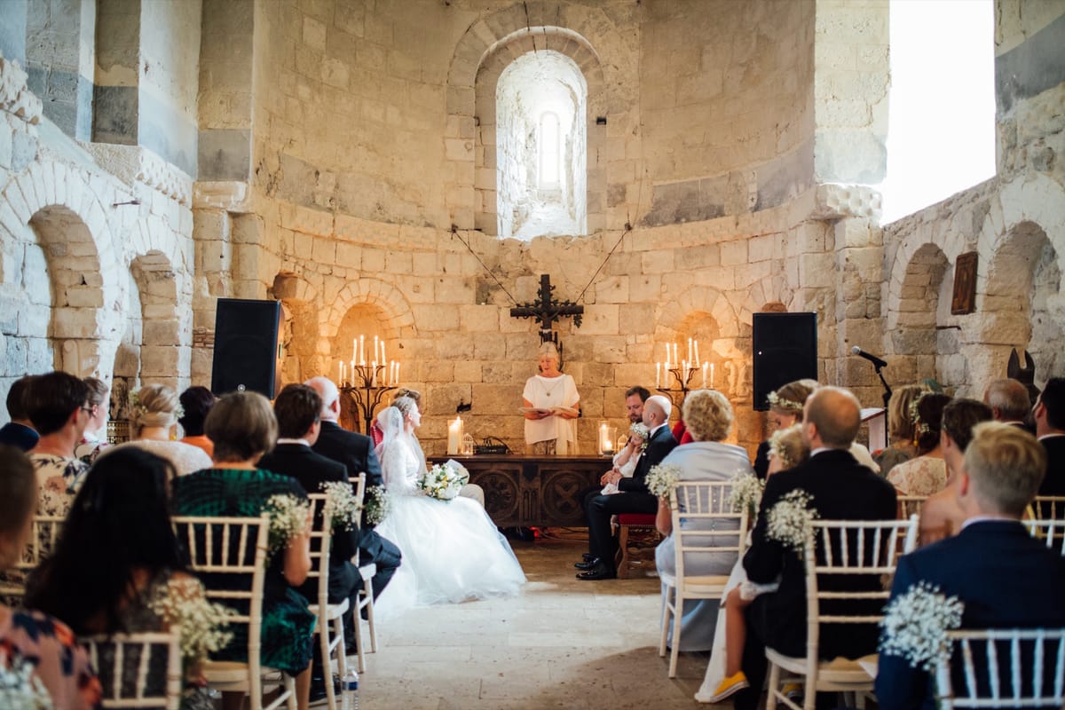 Guests on white chairs watch a ceremony in a vaulted stone chapel lit by candelabras