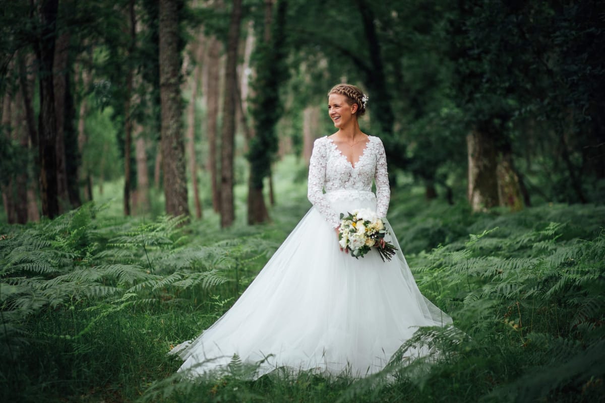 Bride in a long-sleeved lace gown holding a white bouquet among ferns in the estate woodland