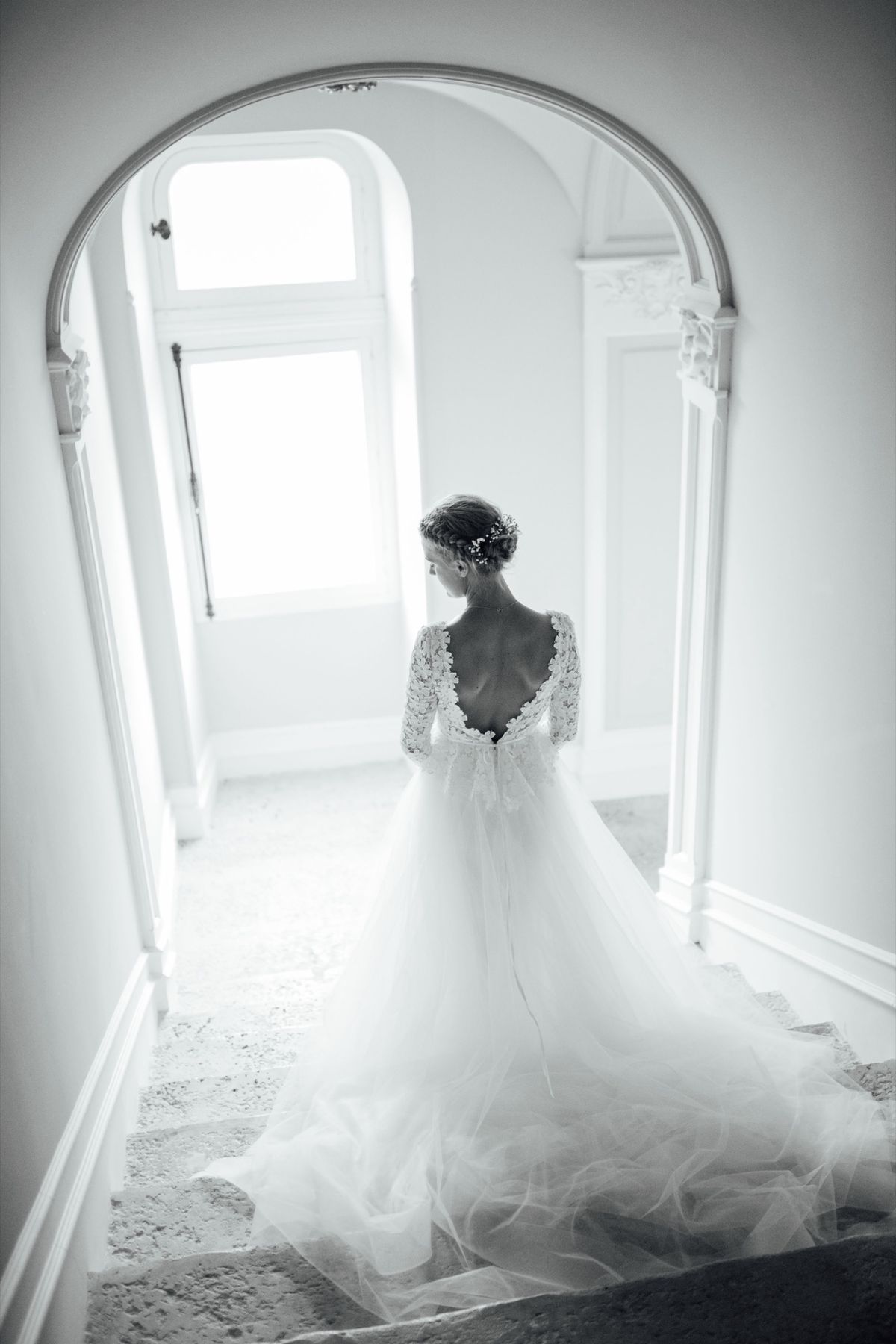 Black and white photo of a bride seen from behind in a backless lace gown, her train spread down the stairs
