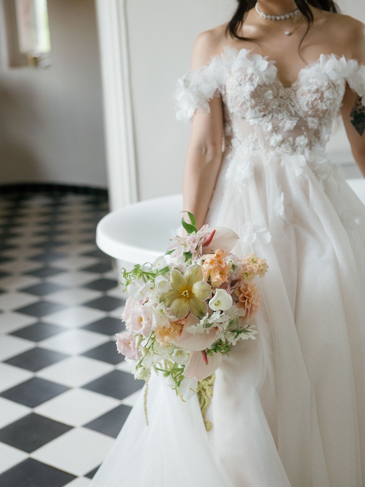 Bride in an off-shoulder lace gown holding a pastel bouquet beside a roll-top bath on chequered tiles