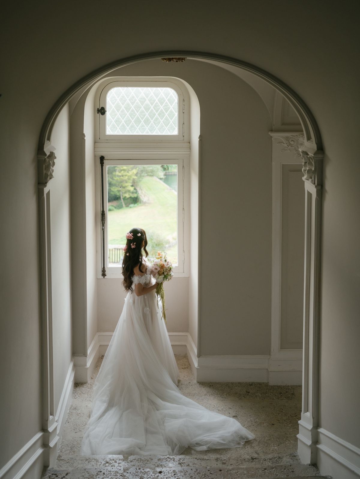 Bride in a long tulle gown holding a bouquet at an arched window, looking out over the lawn