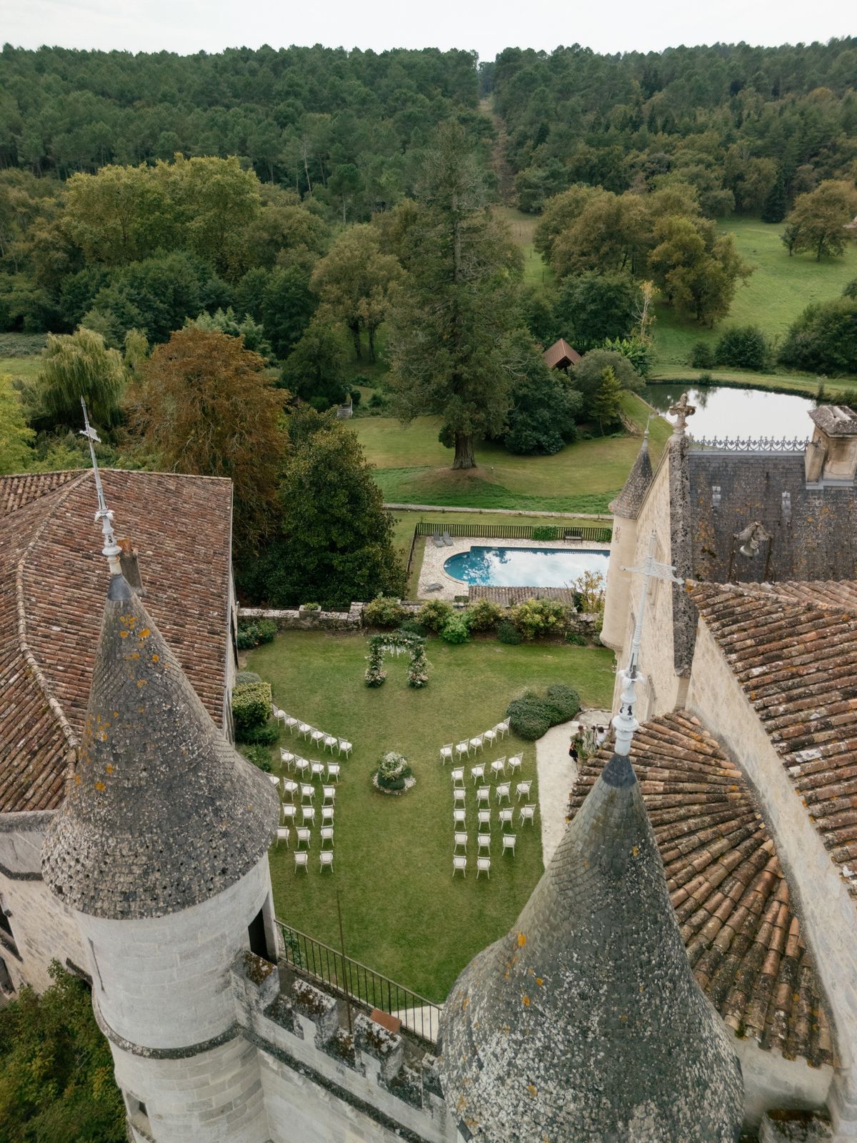 Aerial view of a ceremony set up on the lawn between the towers, with the pool, lake and forest beyond