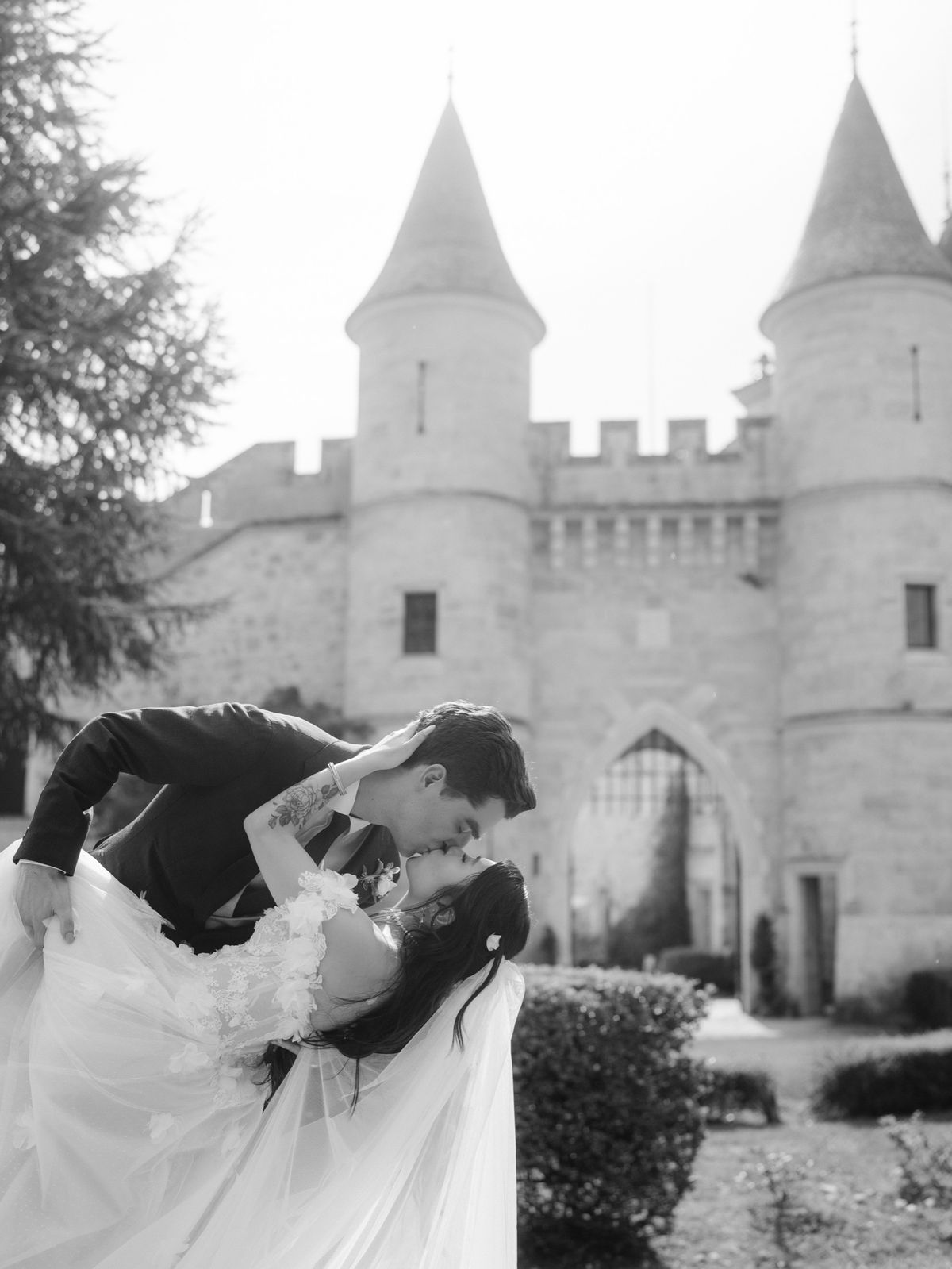 Black and white photo of a groom dipping his bride for a kiss in front of the turreted gatehouse