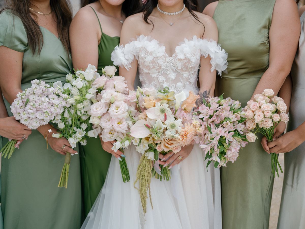 Bride holding a peach and white bouquet between bridesmaids in sage green satin dresses with their own bouquets