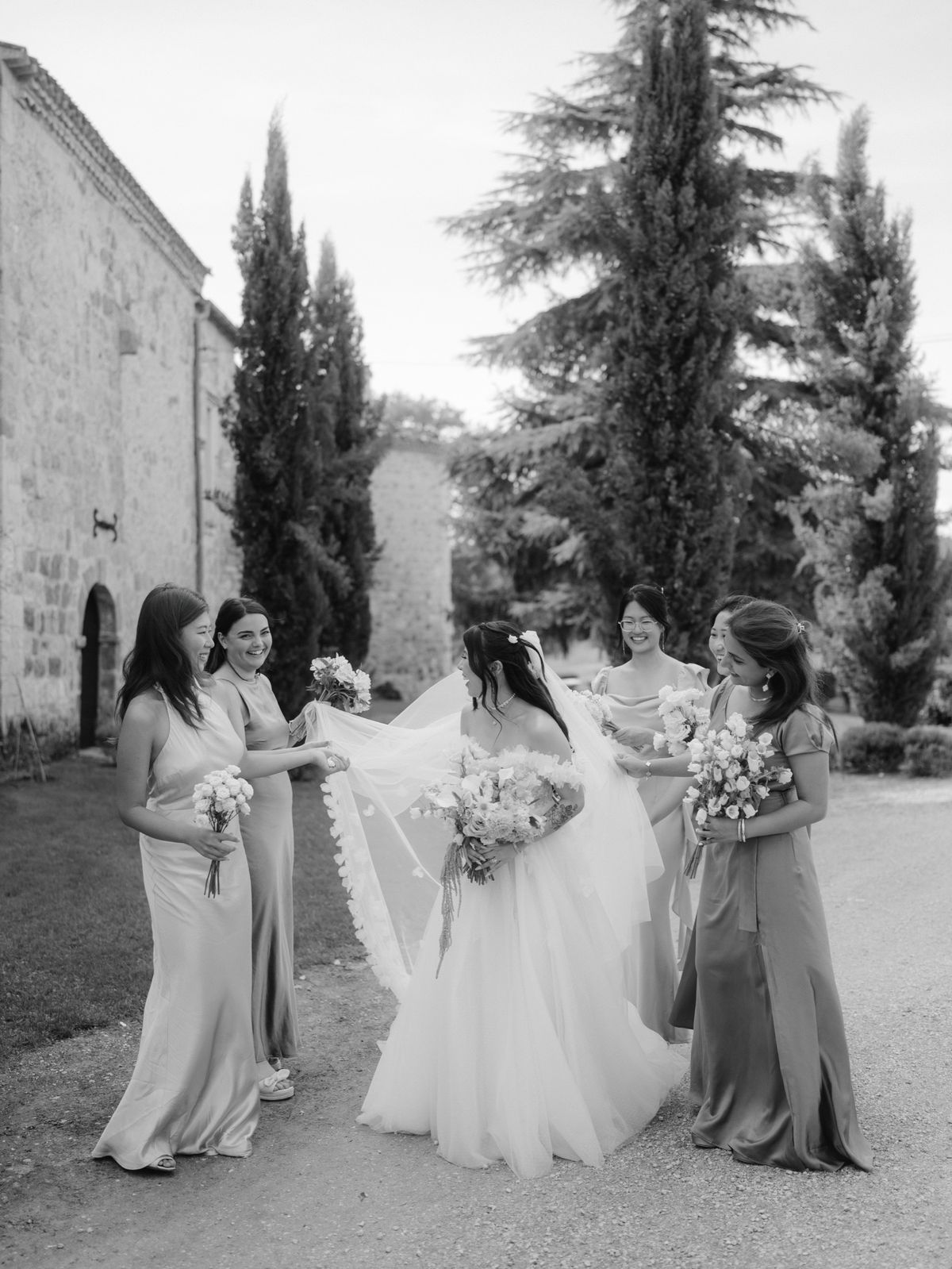 Black and white photo of bridesmaids lifting the bride's long veil on a gravel path lined with cypress trees
