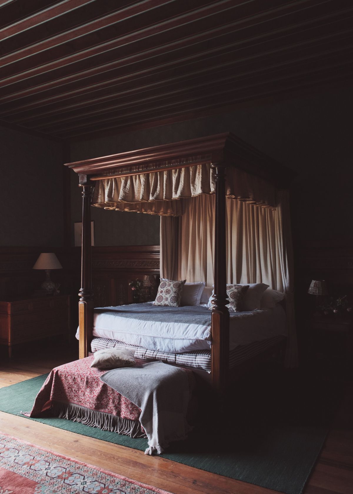 Four-poster bed with cream drapes in a dark bedroom with a striped wooden ceiling