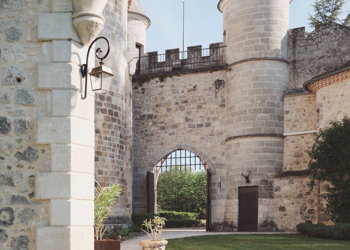Courtyard view of stone towers and a battlemented wall around an arched iron gate, with a mounted stag head