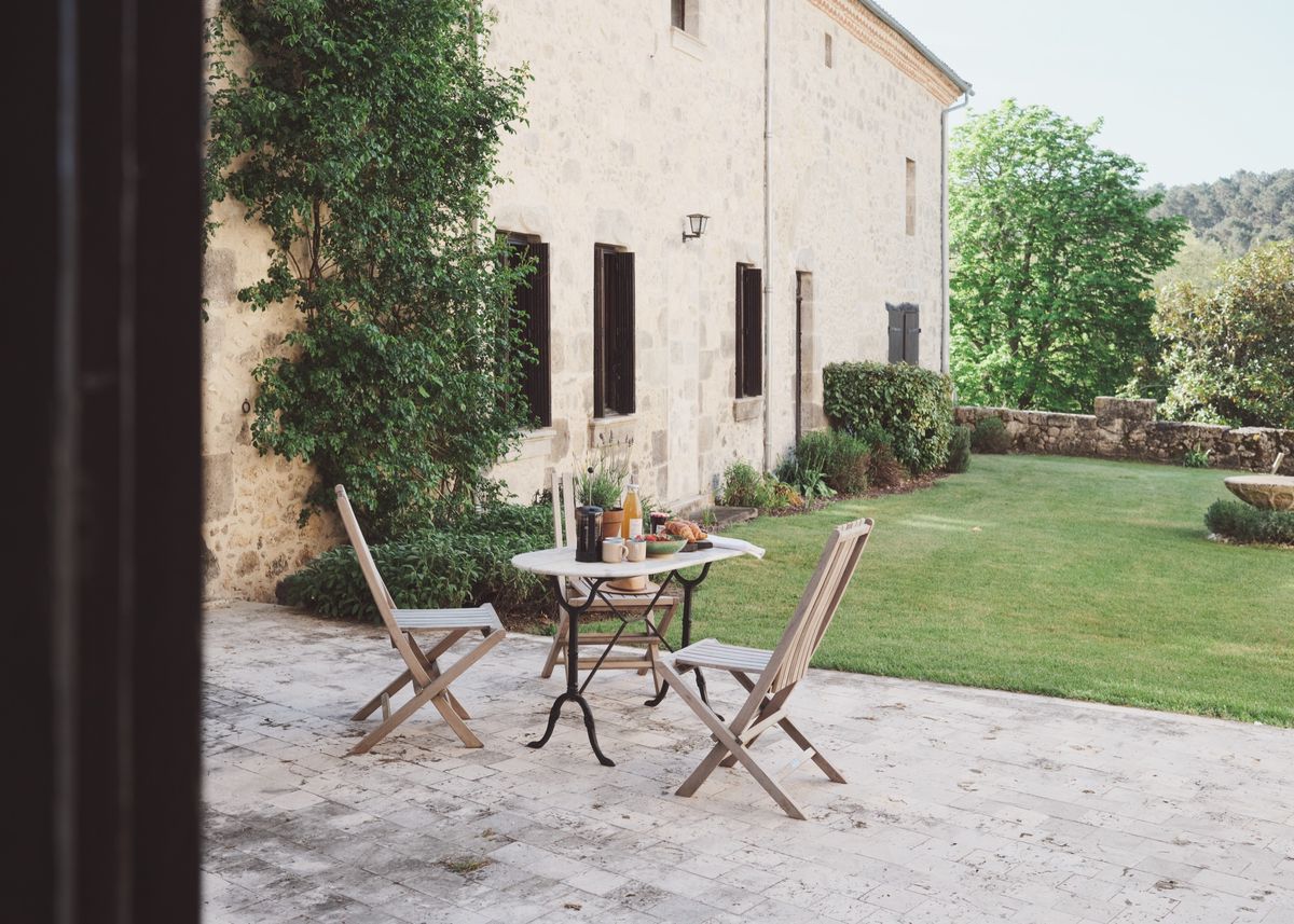 Table laid with breakfast and folding chairs on a stone terrace beside a long stone building and a lawn