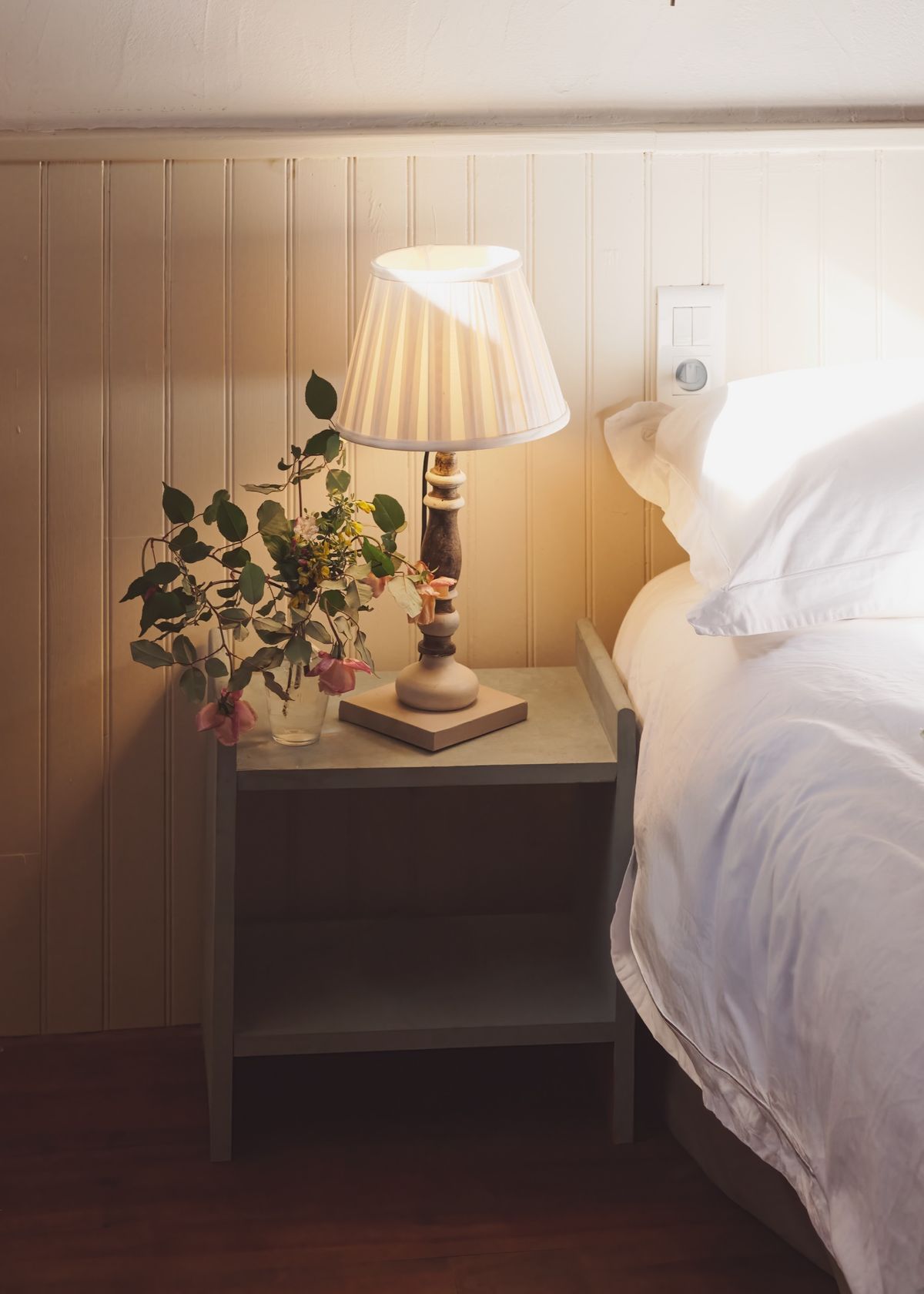 Bedside table with a lamp and a small vase of flowers beside a white bed against panelled walls