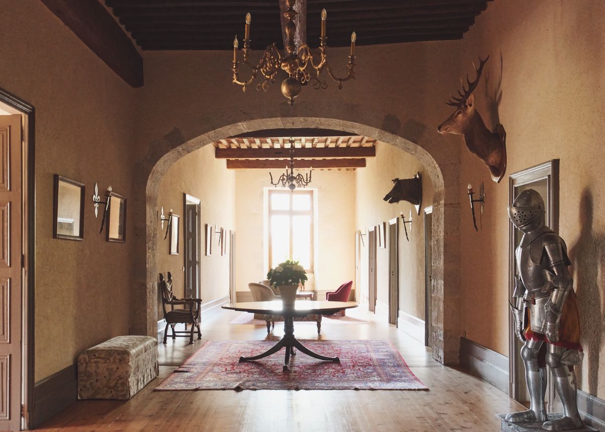 Long hall with a chandelier, a round table on a red rug, stag heads on the walls and a suit of armour