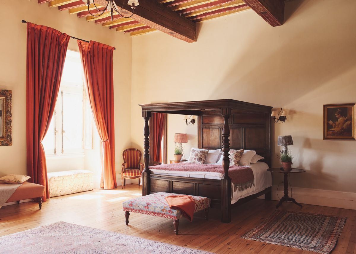 Four-poster bed under a beamed ceiling, with orange curtains at a tall window and a tapestry footstool