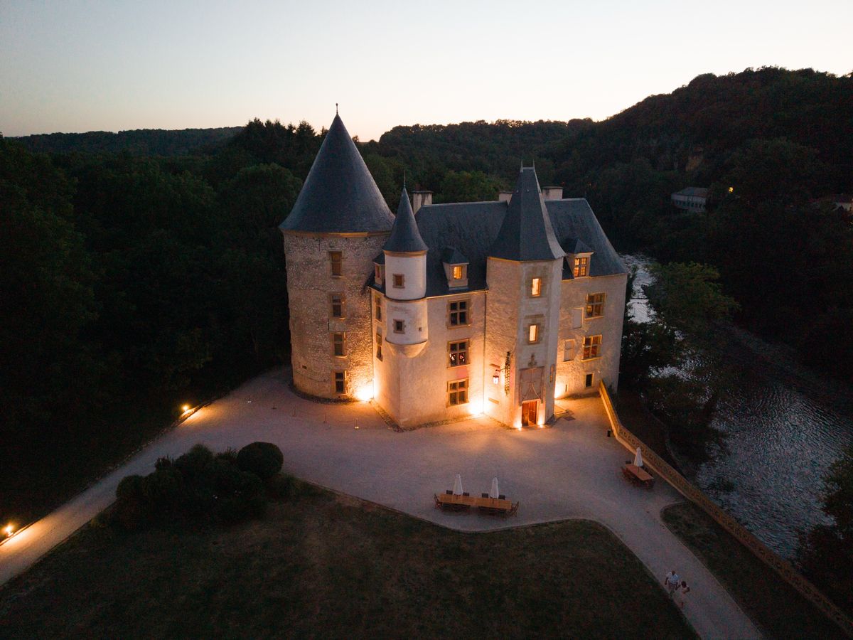 The château lit at dusk, with conical slate towers above a gravel forecourt, a lawn in front and the river on the right