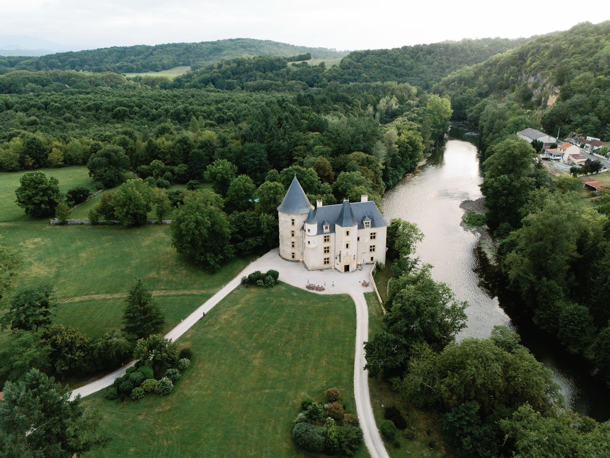 Drone view of the château between open lawns and the river, with a curving gravel drive and wooded hills behind