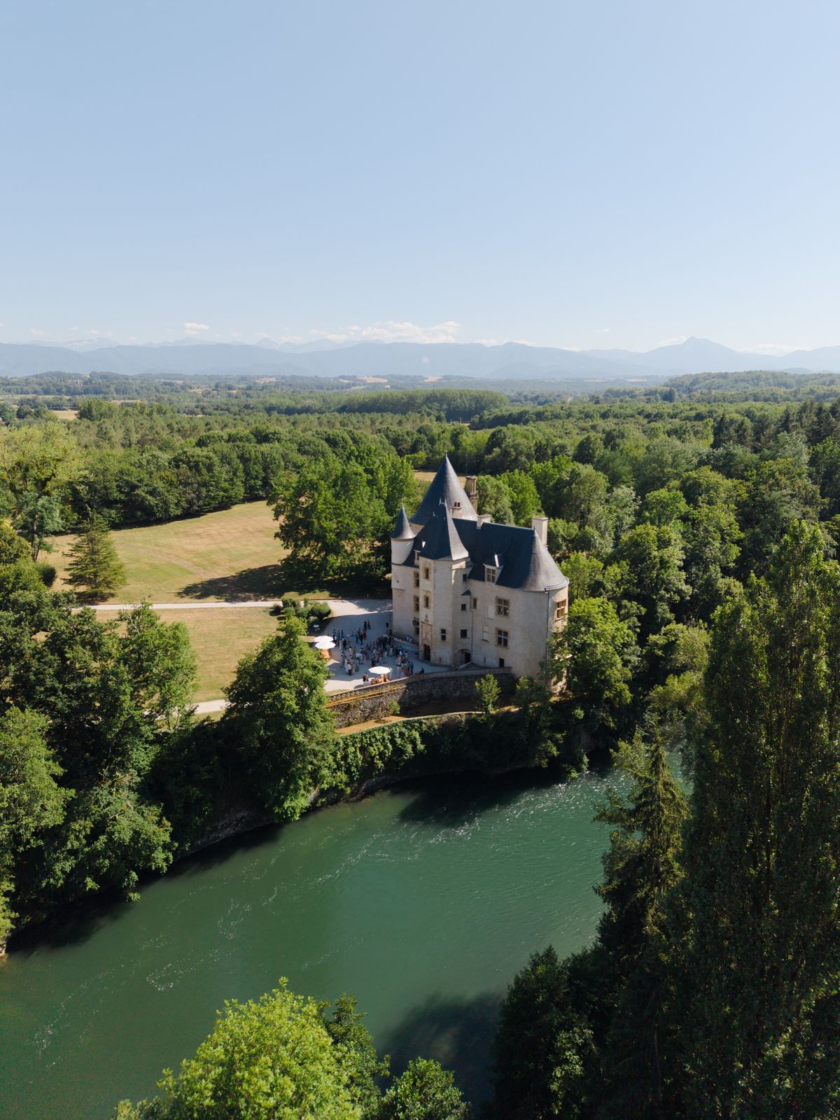 Drone view of the château above a bend of the green river, with guests gathered on its terrace and mountains in the distance