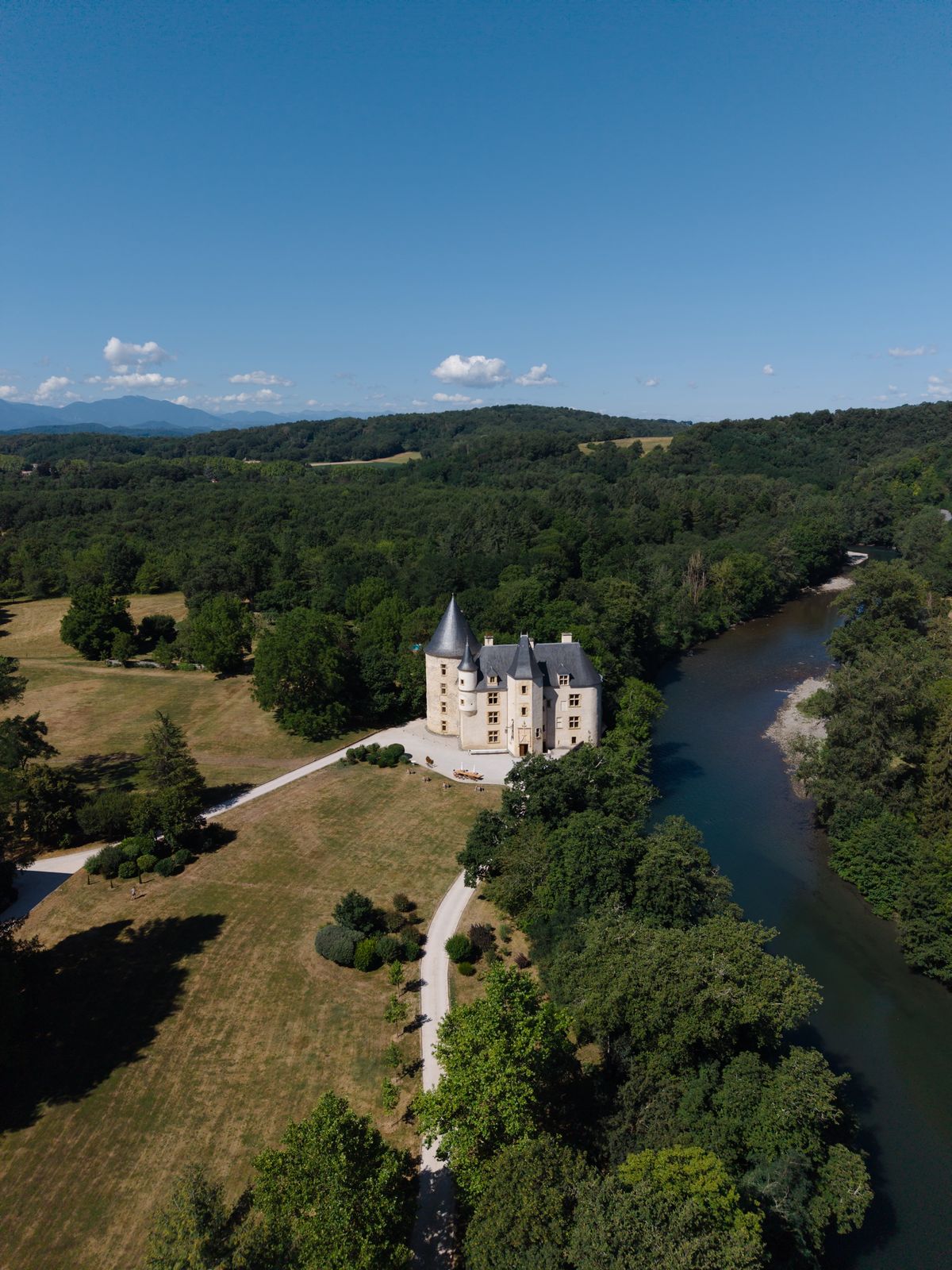 Drone view of the turreted château beside the river, set in parkland with a gravel drive and mountains on the horizon