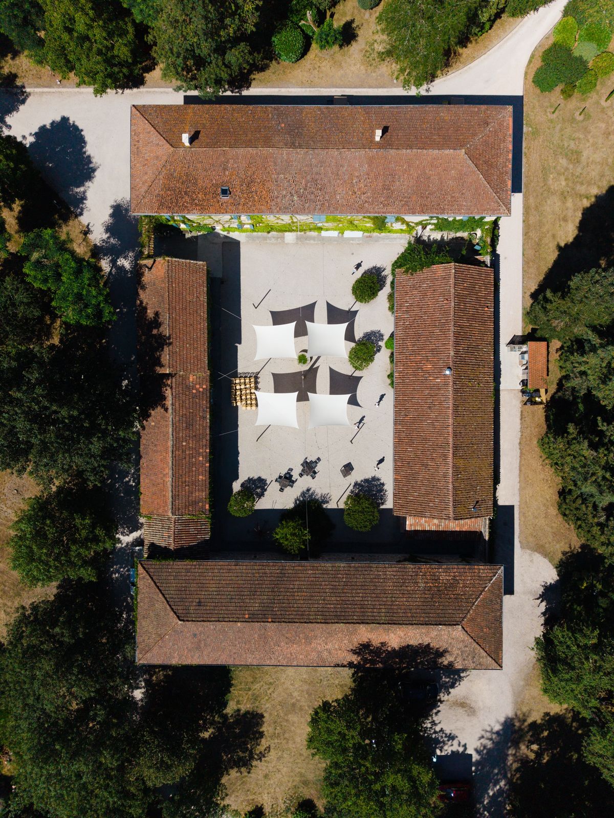 Drone view straight down onto four tiled farm buildings around a gravel courtyard, with white and grey sail shades at its centre