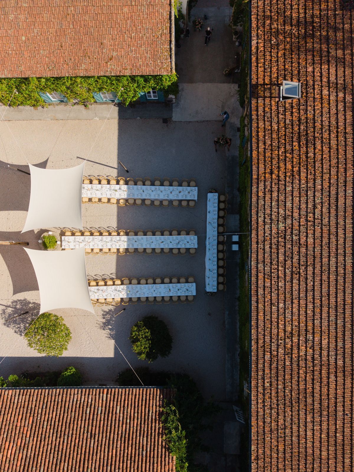 Drone view down onto three long dining tables in a gravel courtyard beside two white sail shades, with a fourth table at the end of the row
