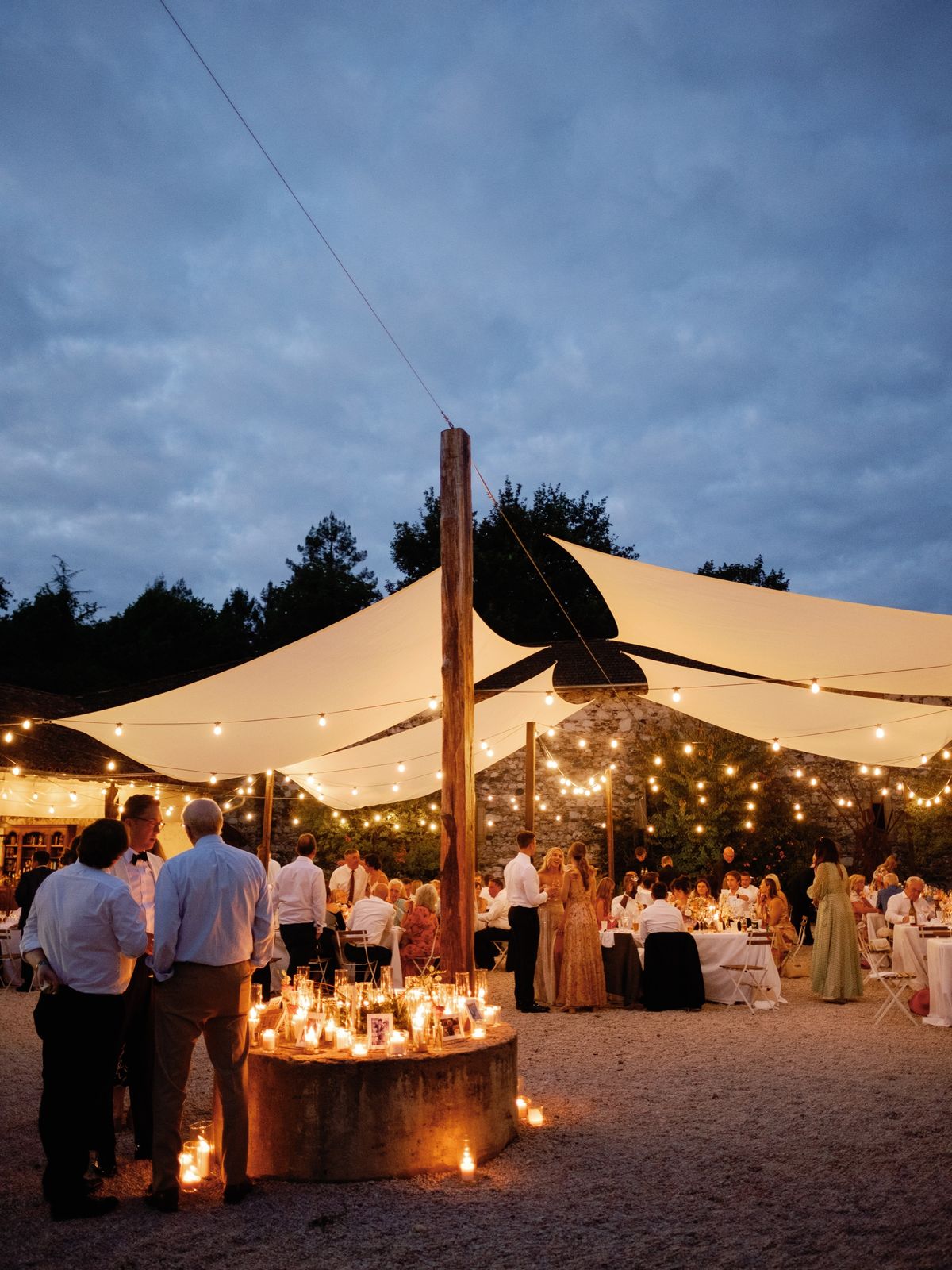 Evening reception in the courtyard under white sail shades and festoon lights, with guests standing around a round stone base covered in candles