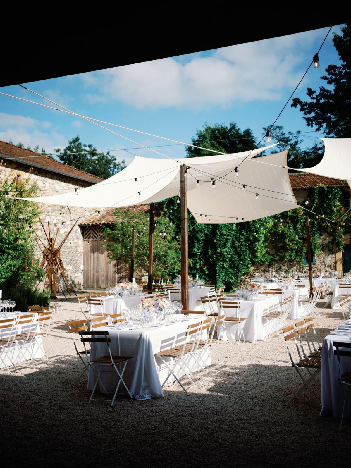 Courtyard laid for a daytime reception, with white-clothed tables and wooden folding chairs under white sail shades and festoon lights, against ivy-covered stone barns