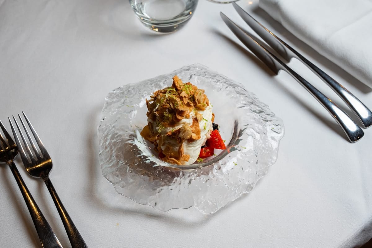 A cream dessert topped with toasted almond flakes and lime zest, with strawberries and berries, in a textured glass bowl