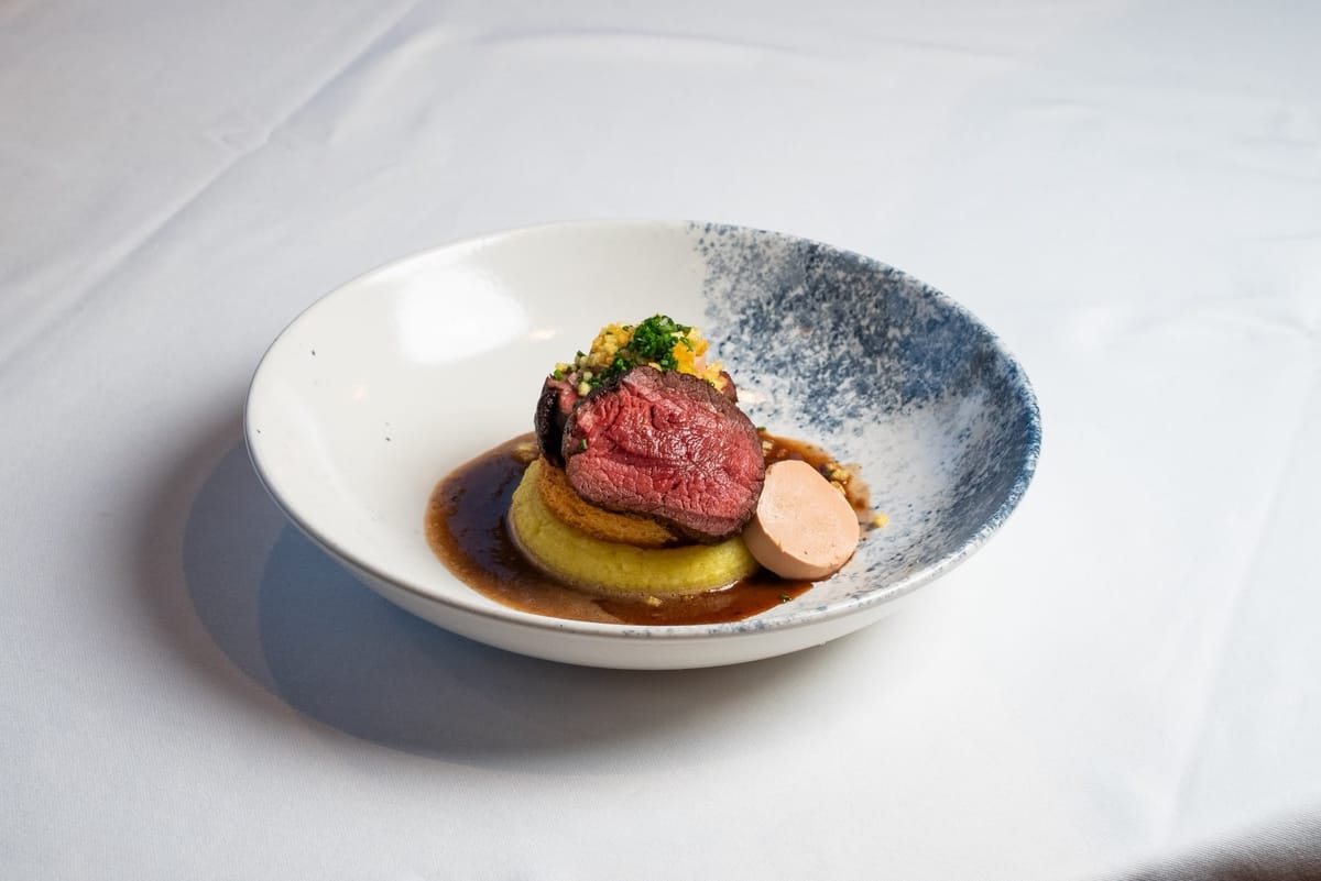 Seared beef fillet and foie gras on toasted bread over potato purée, with jus, in a half blue-speckled bowl