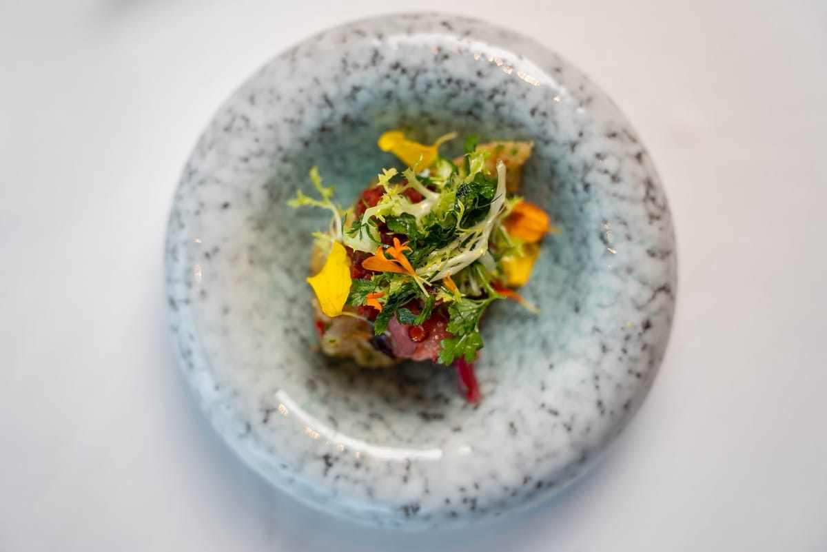 Octopus and pickled onion under frisée, herbs and yellow and orange edible flowers, seen from above in a speckled bowl