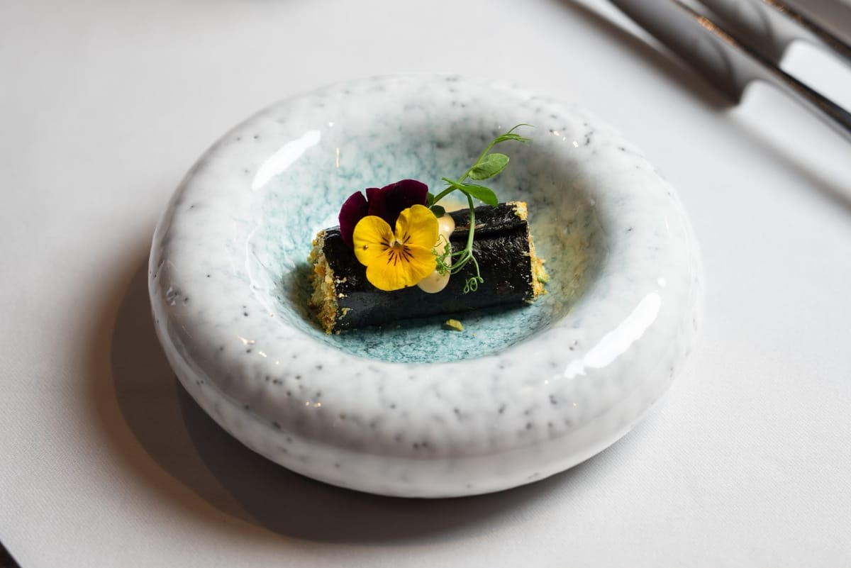 A dark rolled starter topped with a yellow pansy and pea shoots, in a turquoise-glazed bowl