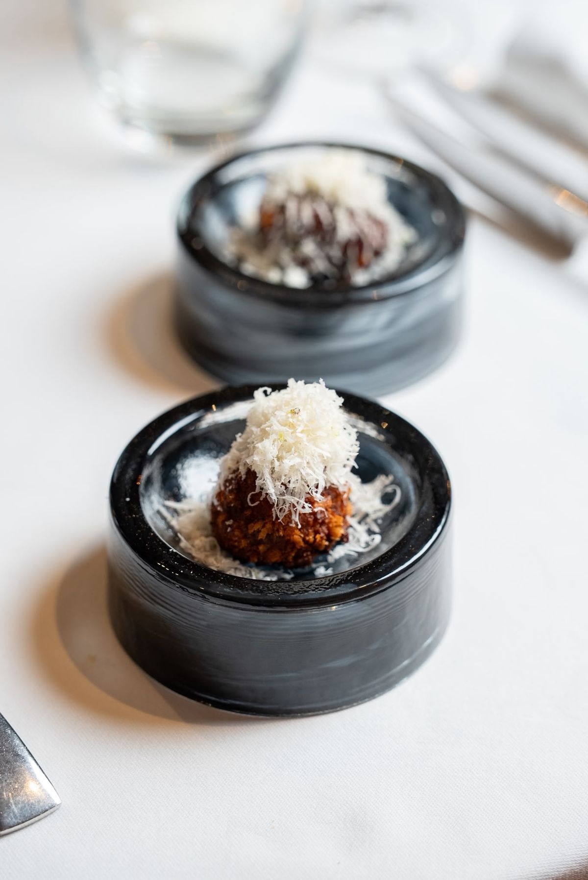 Two fried croquettes topped with finely grated white shavings, on small round black glass dishes