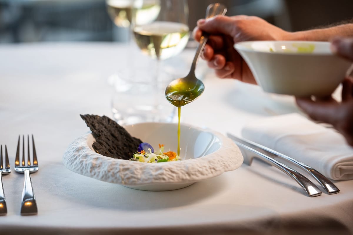 A hand spoons green herb oil over a starter with edible flowers and a black crisp, at a laid table