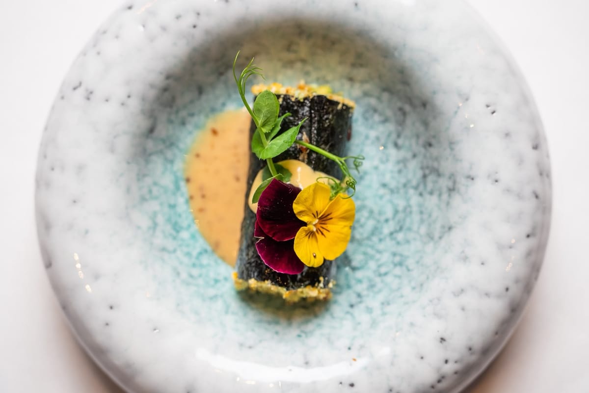 A rolled starter with a pansy and pea shoots over a pale sauce, seen from above in a turquoise bowl