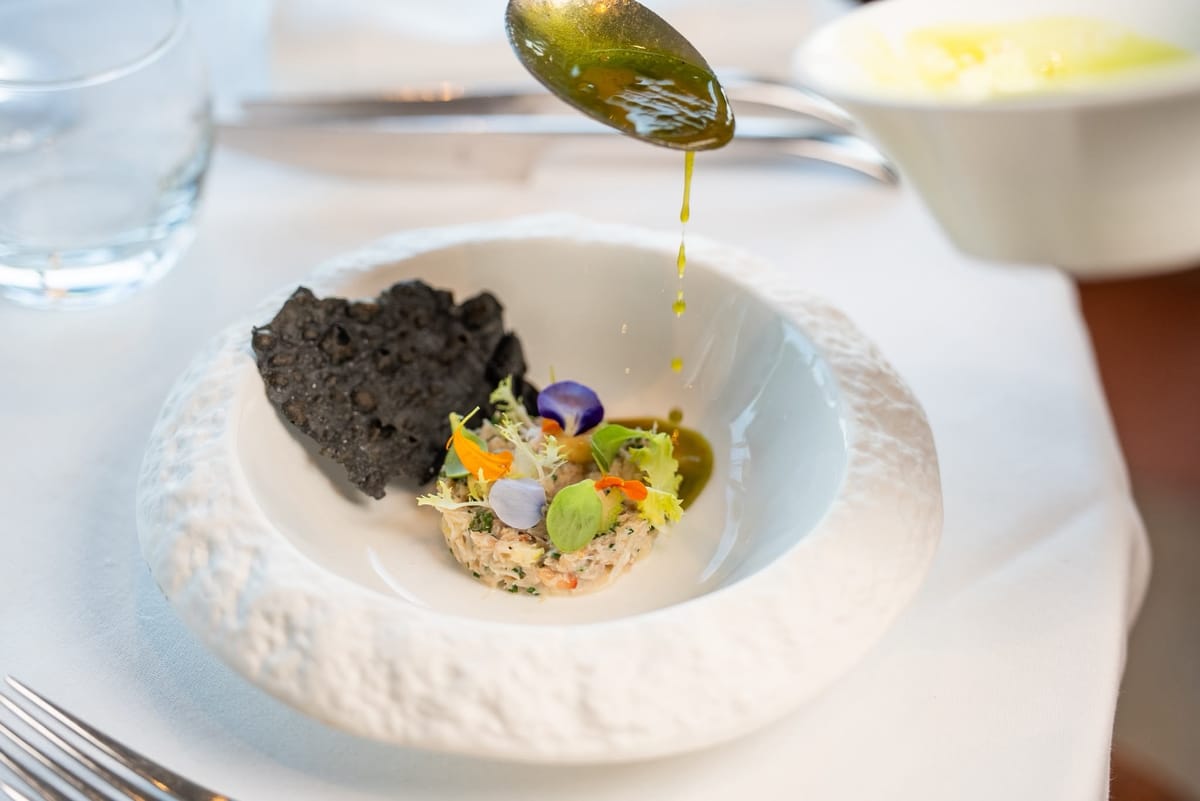 Herb oil poured from a spoon over a starter with edible flowers and a black crisp