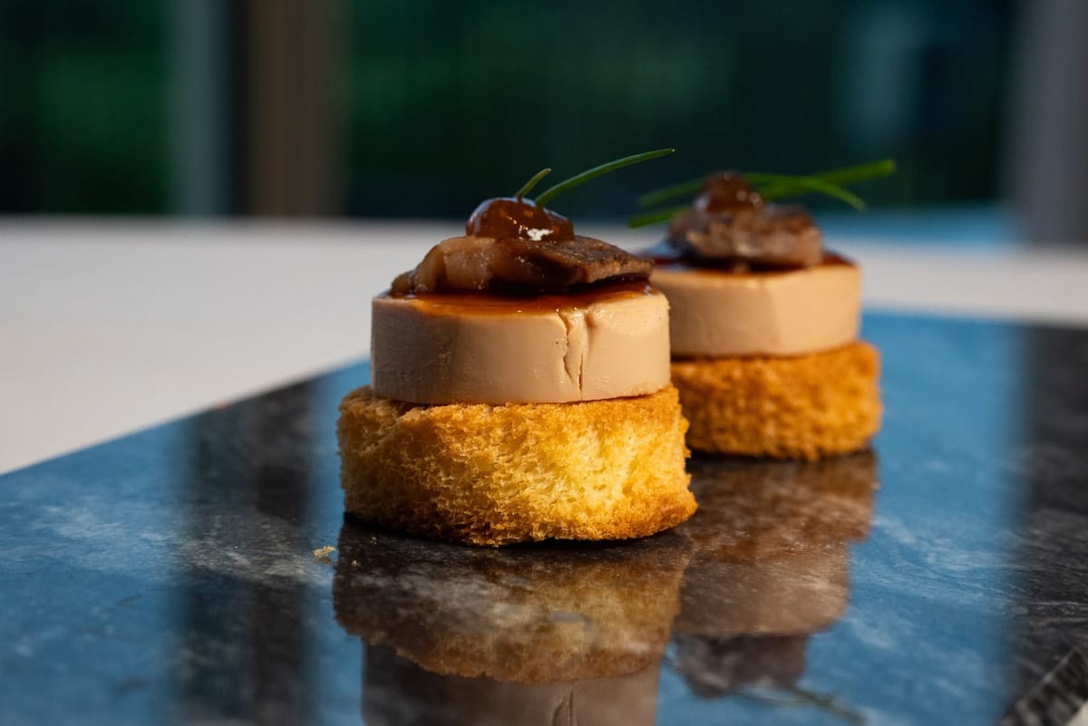 Two foie gras rounds on toasted brioche, topped with a glazed garnish, chutney and chives, on dark marble