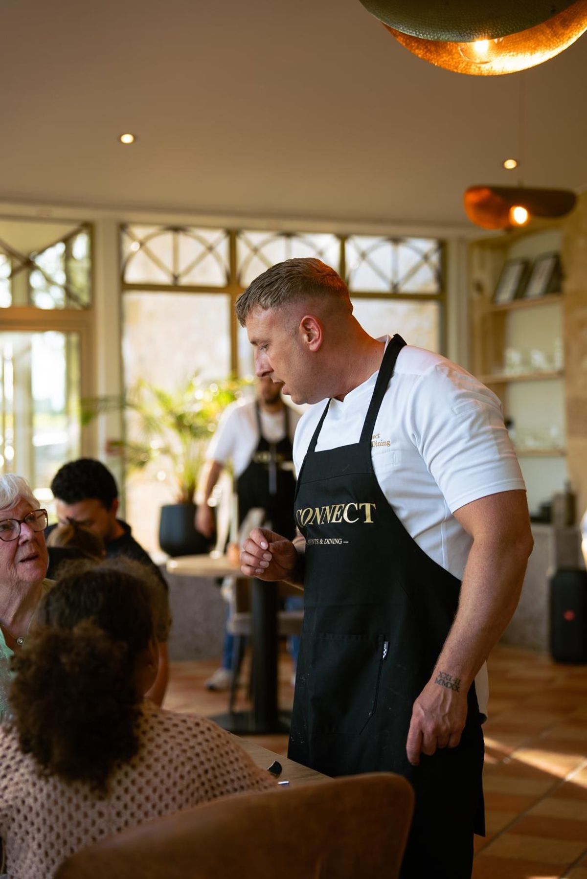 A staff member in a black apron talks with seated guests in a sunlit dining room, a colleague behind