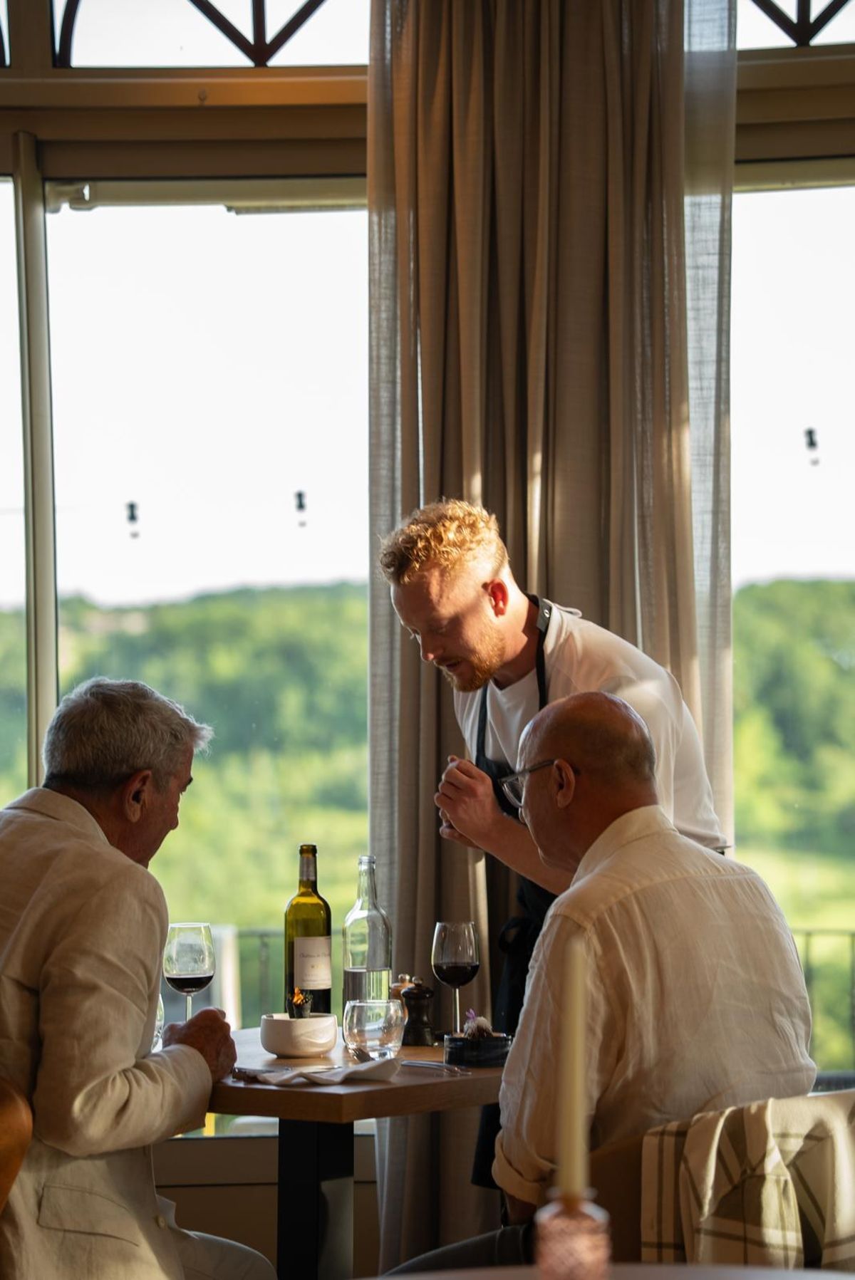 A staff member leaning over two men at a window table, with wooded hills beyond