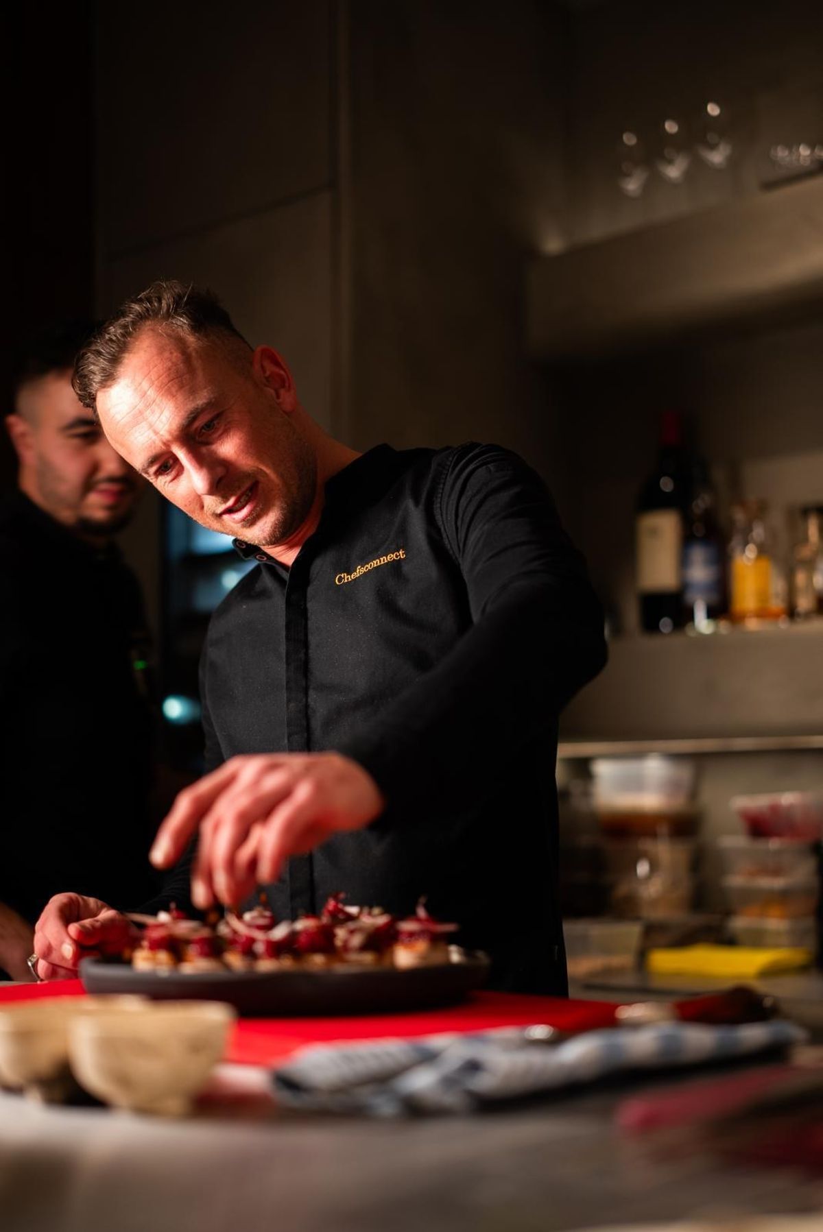 A chef in a black jacket plating a tray of canapés in the kitchen, a colleague behind him