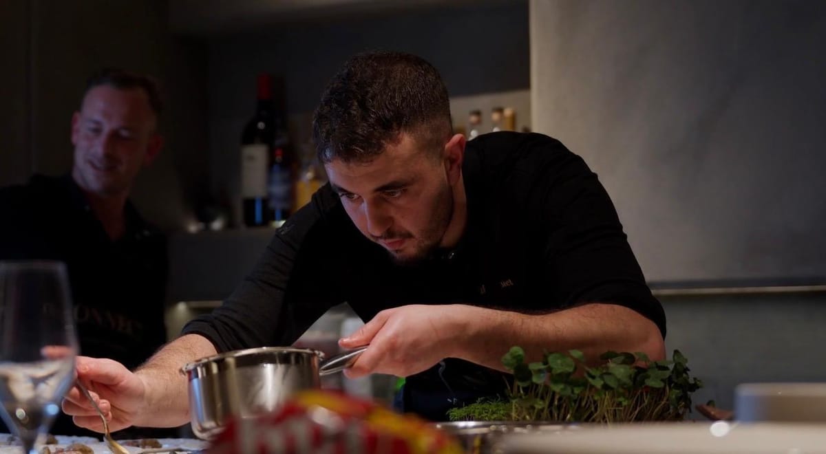 A chef leaning in to finish a plate with a small pan, a colleague smiling in the background