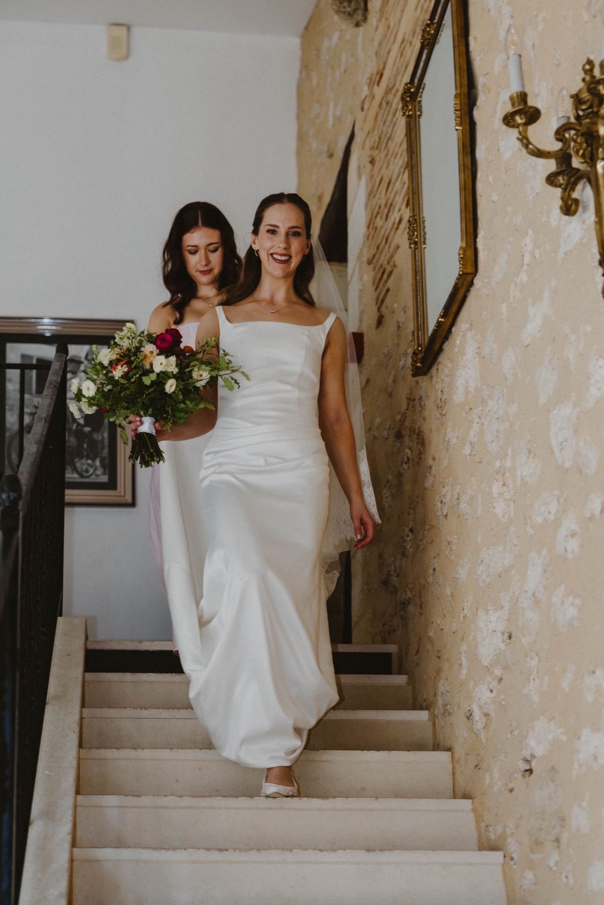 A smiling bride holding a red, cream and peach bouquet walks down a stone staircase, a woman in pale pink behind her