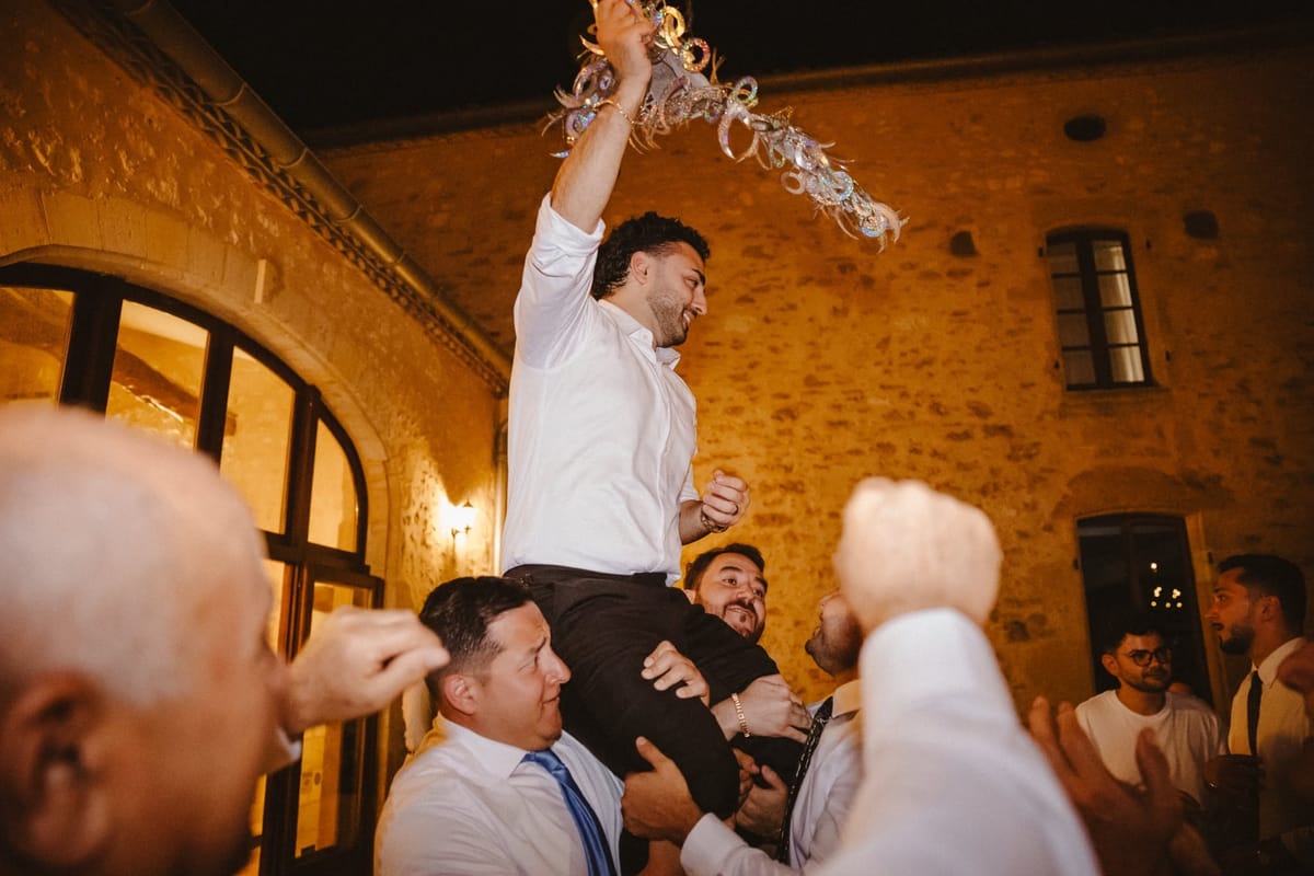 Guests lift a man in a white shirt onto their shoulders as he waves a sparkly streamer in the lit stone courtyard at night