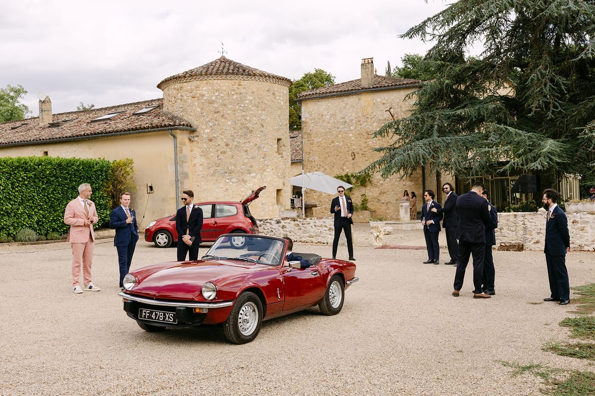 Men in suits gather around a red vintage convertible on the gravel courtyard before the stone château and its round tower