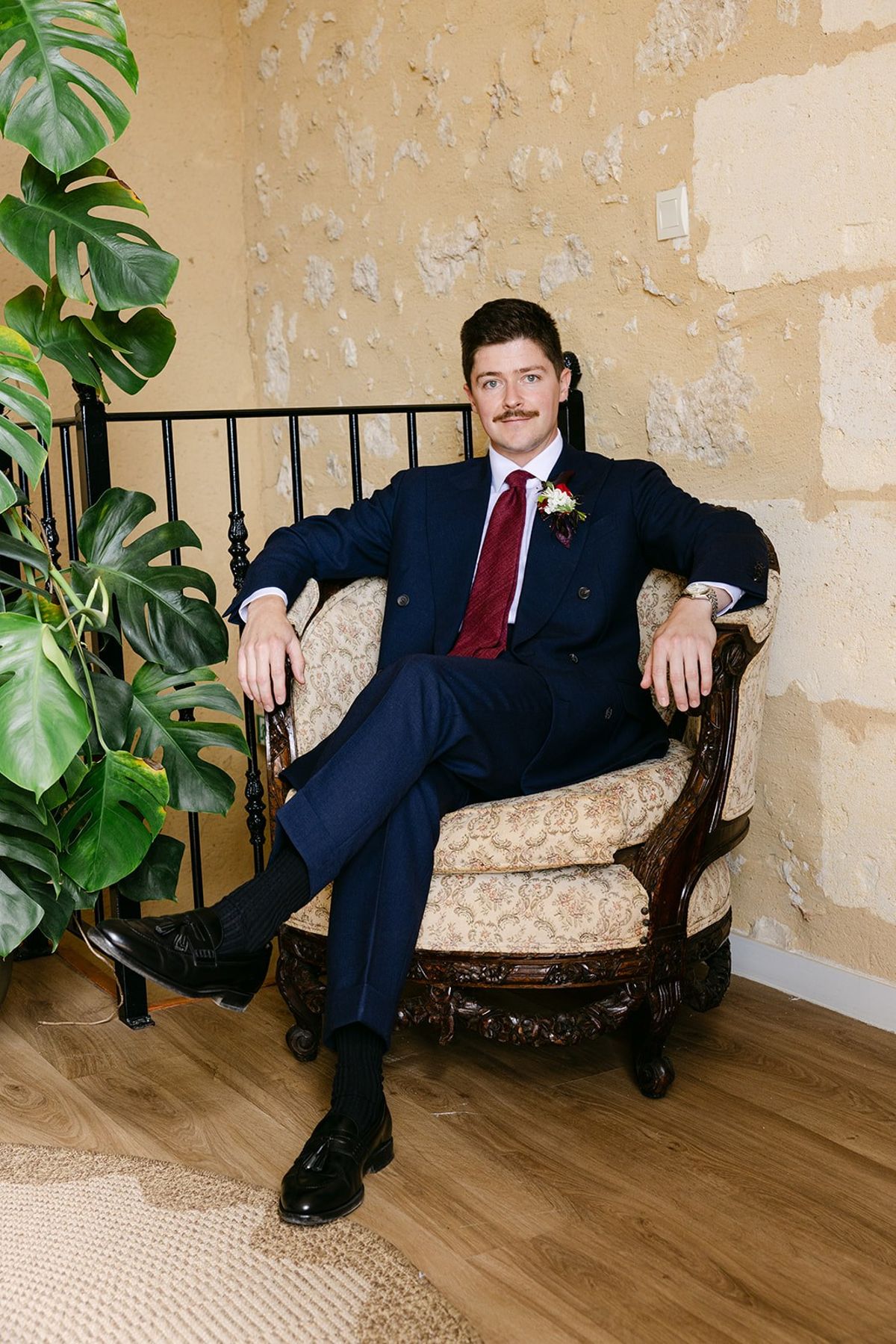 Man in a navy suit and burgundy tie seated in an antique armchair by an iron stair rail and pale stone wall