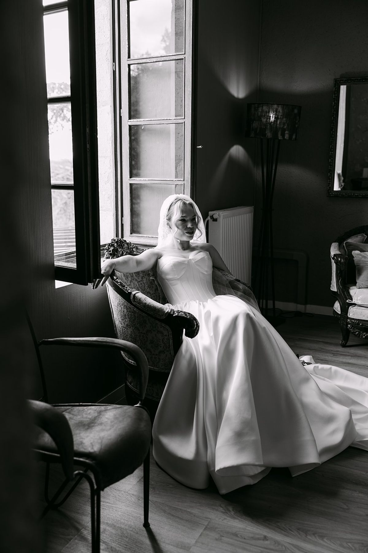 Bride in a strapless gown and veil seated in an armchair beside a tall open window, in black and white