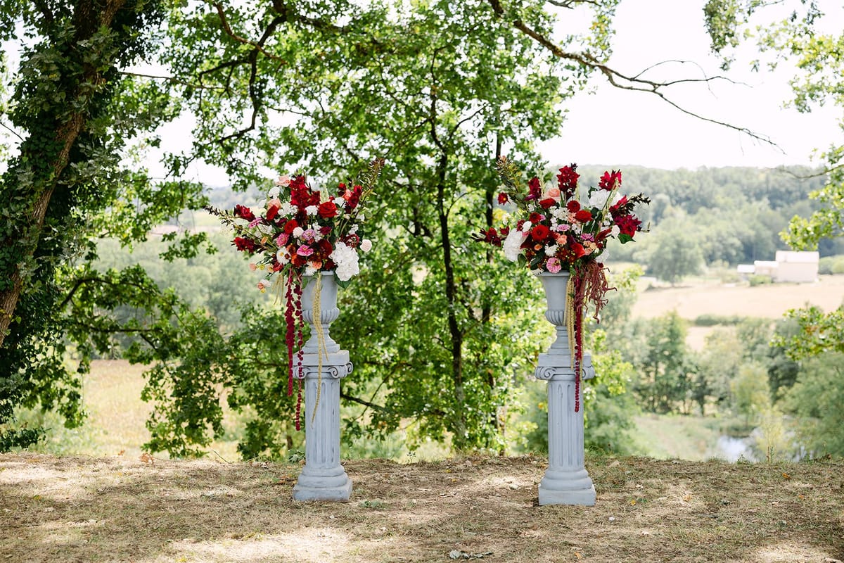 Two grey columns topped with red and white flower urns under trees, with fields and wooded hills beyond