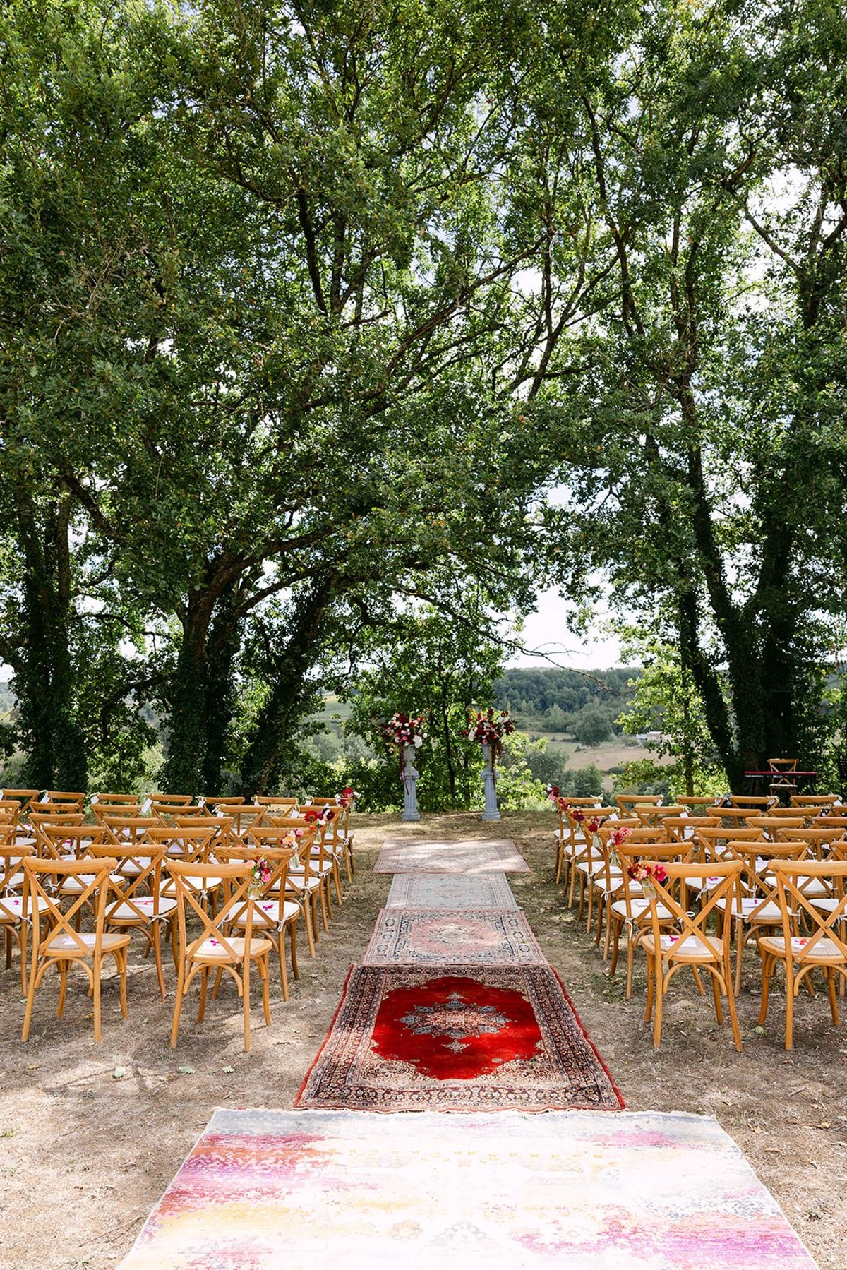 Rows of wooden cross-back chairs either side of a rug-lined aisle under tall trees, flower columns and hills beyond