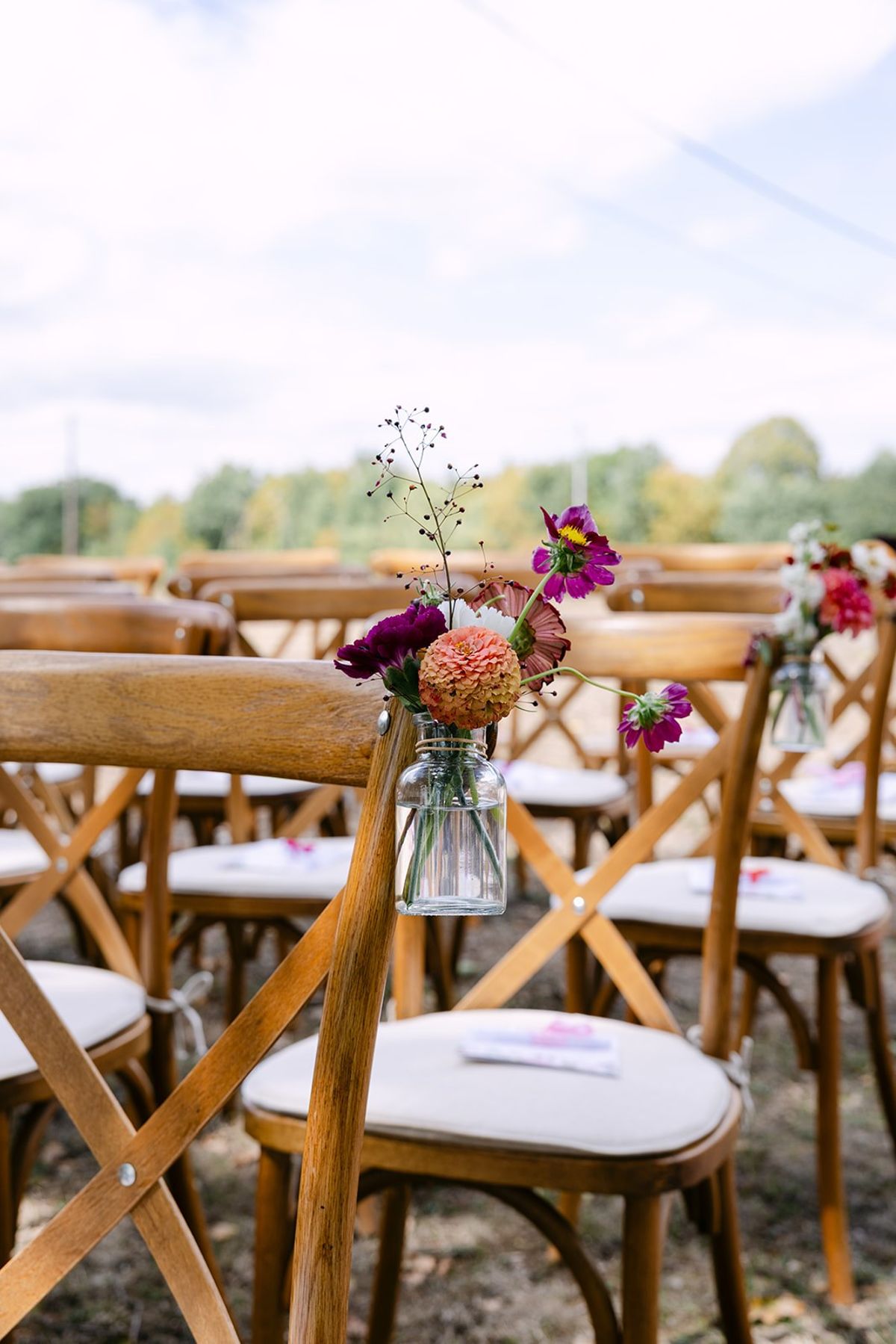 Small glass jar of pink, orange and purple flowers hung on a wooden cross-back chair, rows of cushioned chairs behind