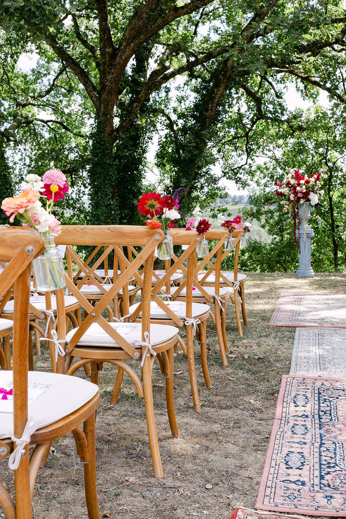 Cross-back chairs with jars of pink and red flowers beside a rug-lined aisle under large trees, a flower column ahead