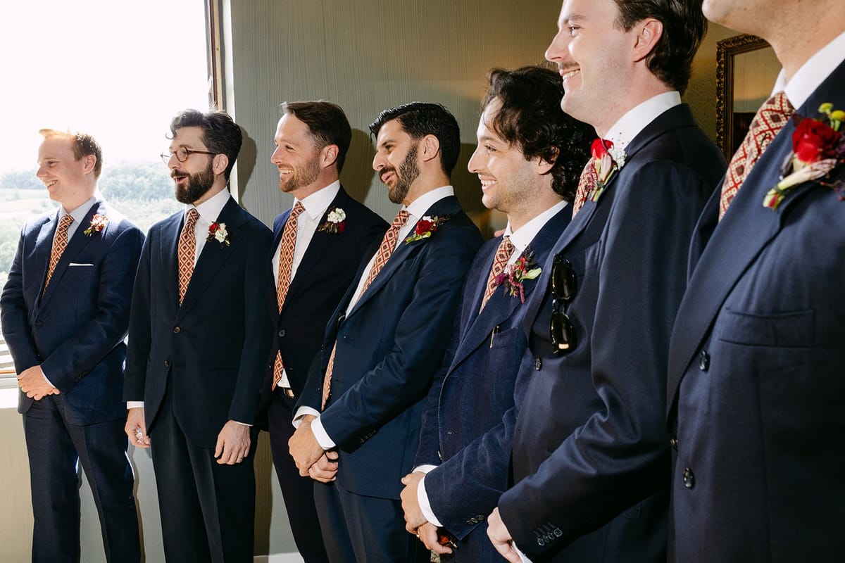A row of men in navy suits and patterned ties with red buttonholes stand by a window overlooking green countryside