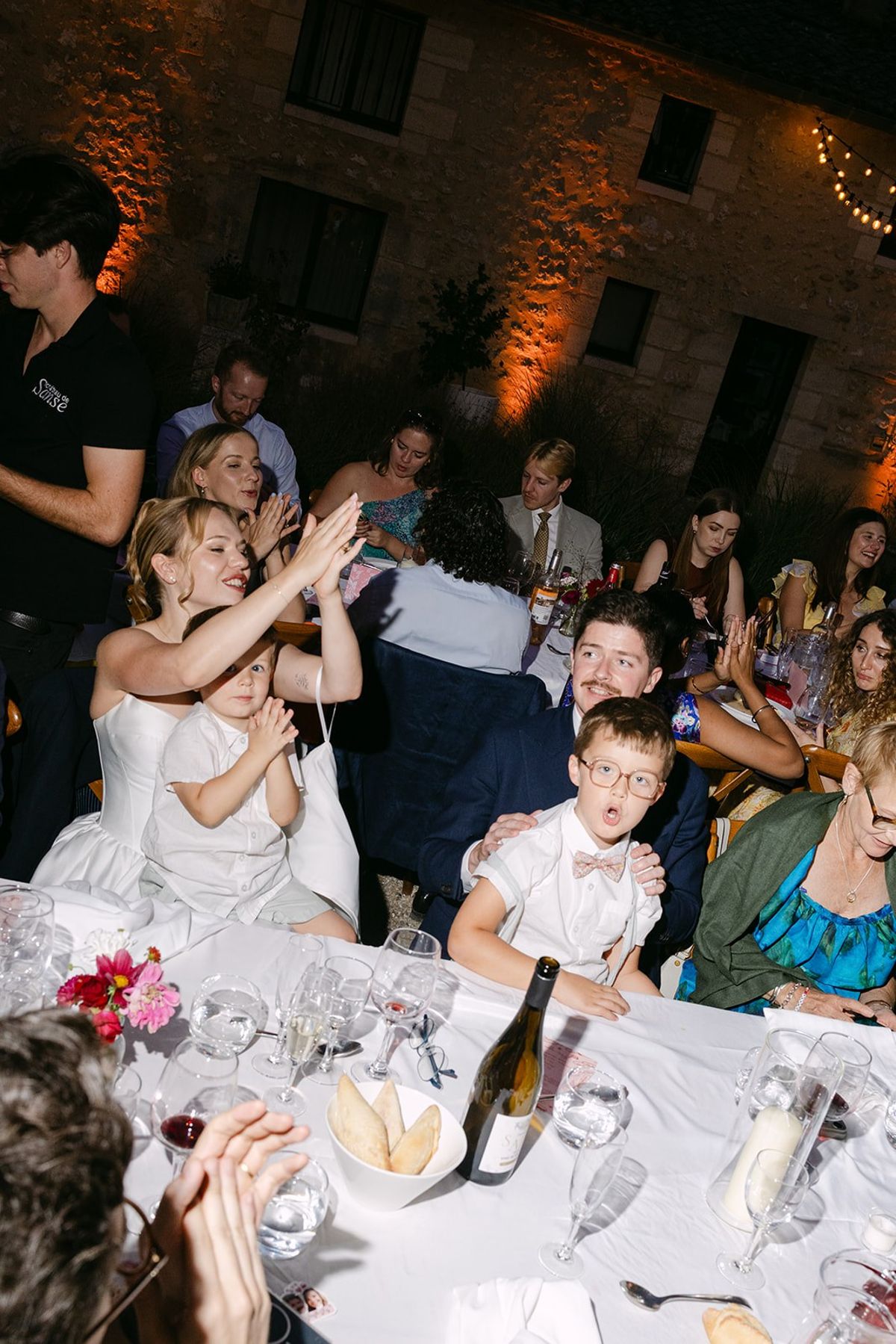 Bride and guests clap at a white-clothed table at night, the stone château wall uplit in amber behind