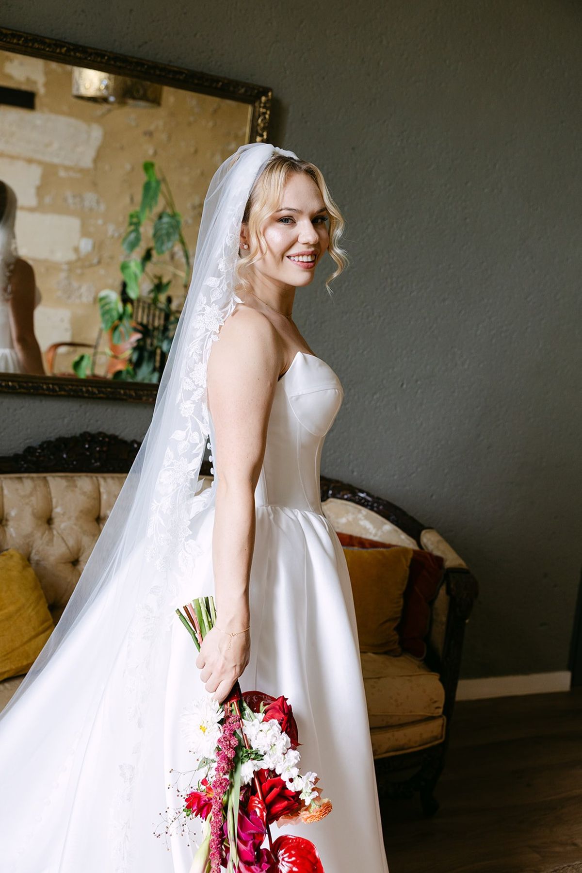 Bride in a strapless gown and long lace-edged veil holding a red and white bouquet beside a gilt mirror and tufted sofa