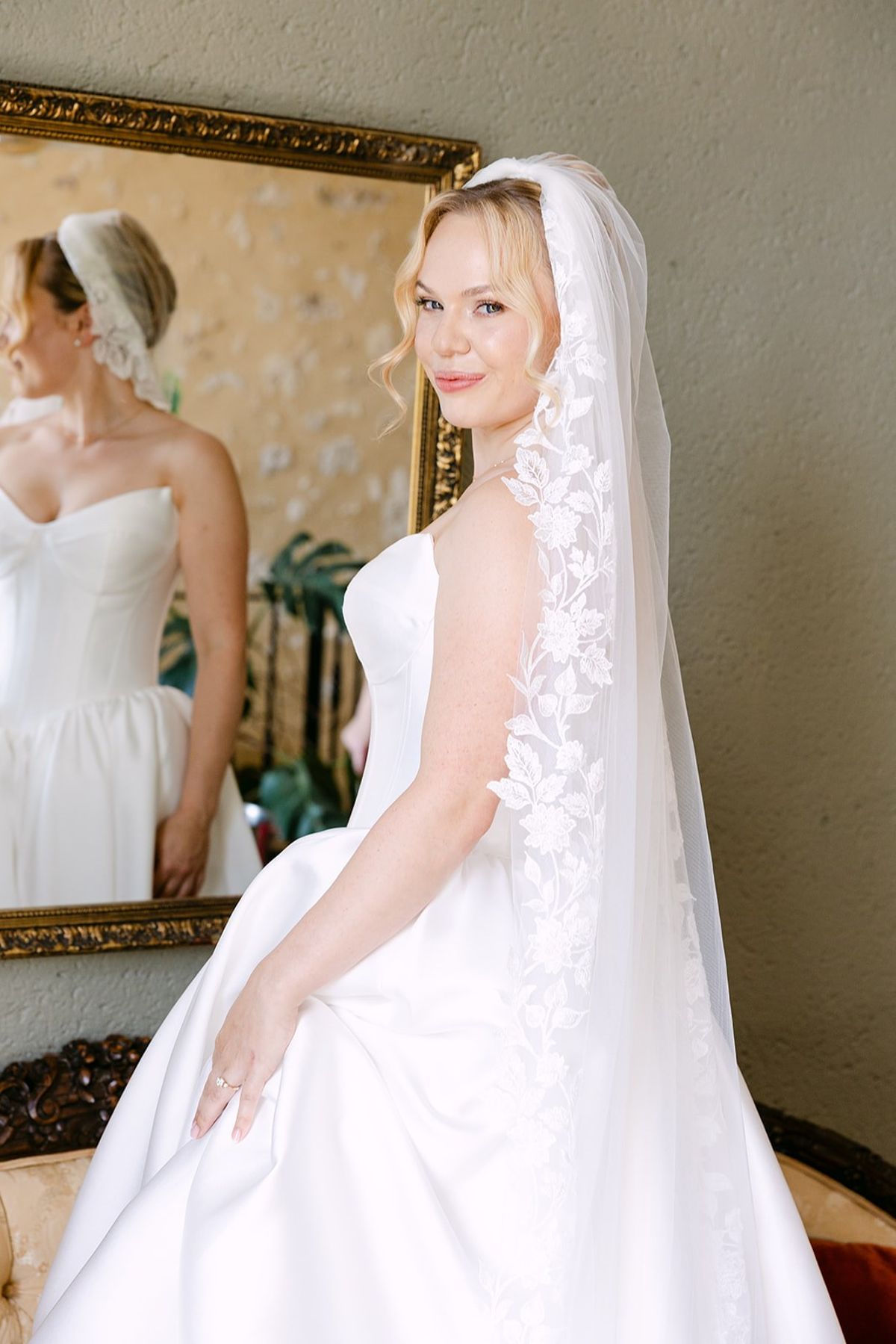 Bride in a strapless gown and lace-edged veil glances over her shoulder in front of a gilt-framed mirror