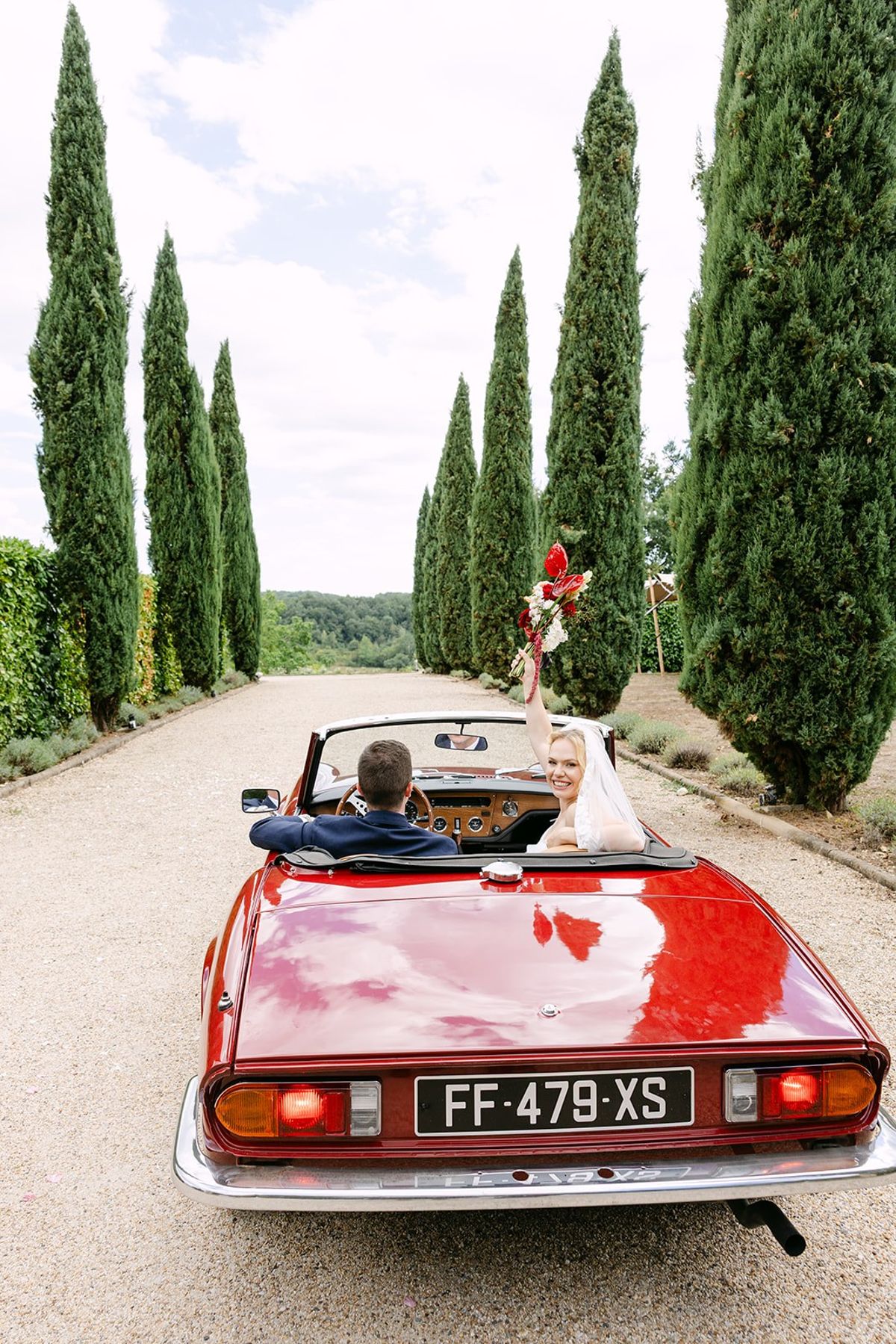 Bride raises her bouquet from a red convertible driving down the cypress-lined gravel drive